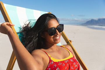 Wall Mural - Smiling mixed race woman on beach holiday sitting in deckchair sunbathing