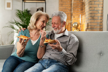 Wall Mural - Cheerful husband and wife sitting on sofa at home. Happy senior woman and man eating pizza while watching a movie.