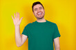 © Jihan - young Caucasian man wearing green T-shirt against yellow wall Waiving saying hello happy and smiling, friendly welcome gesture.