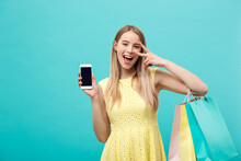 Portrait Young Attractive Woman With Shopping Bags Shows The Phone's Screen Directly To The Camera. Isolated On Blue Background