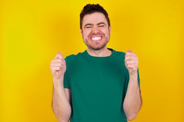 Sticker - young Caucasian man wearing green T-shirt against yellow wall being excited for success with raised arms and closed eyes celebrating victory. Winner concept.