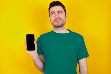 Wall Mural - young handsome caucasian man wearing green t-shirt against yellow background holds new mobile phone and looks mysterious aside shows blank display of modern cellular