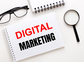 Fototapete - DIGITAL MARKETING is written in a white notebook on a light background near the notebook, black-framed glasses and a magnifying glass.