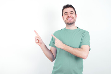 Wall Mural - young handsome caucasian man wearing green t-shirt against white background with positive expression, indicates with fore finger at blank copy space for your promotional text or advertisement.