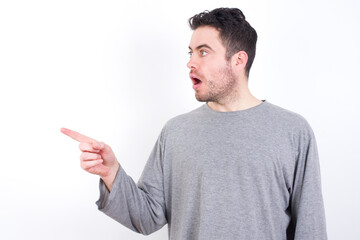 Wall Mural - Stunned young handsome caucasian bearded man wearing pyjama over white wall with greatly surprised expression points away on copy space, indicates something