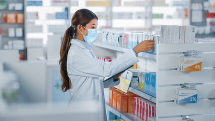 Pharmacy Drugstore: Portrait of Beautiful Pharmacist Wearing Face Mask Uses Digital Tablet Computer, Looks at Camera and Smiles Charmingly, Behind Her Shelves Full of Medicine Packages. Side View Shot