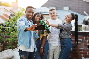 Wall Mural - Cheerful mixed race friends toasting with alcoholic drinks and taking selfie on modern smartphone. Young male and female people having party time with fun.