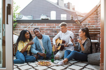 Wall Mural - Four multiracial friends sitting on open terrace, eating tasty pizza and drinking alcohol. Handsome man playing guitar and singing. Party time.