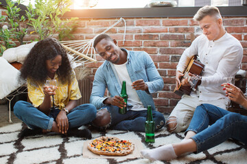 Wall Mural - Foup happy multicultural people in stylish clothes enjoying party time on open terrace. Men and women eating pizza, drinking alcohol and playing guitar. Concept of friendship, fun and relaxation.