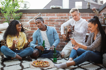 Wall Mural - Happy diverse friends sitting together on open terrace with tasty pizza and alcoholic drinks. Young people playing guitar and singing during partytime.