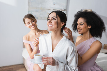 Wall Mural - cheerful woman holding coffee cup near interracial friends preparing her for wedding.