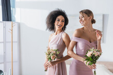 joyful multicultural bridesmaids holding wedding bouquets in bedroom.