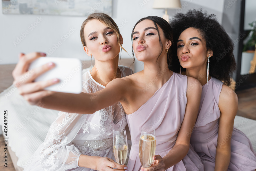 pretty bride with multicultural bridesmaids blowing air kisses while taking selfie on blurred foreground. - obrazy, fototapety, plakaty 
