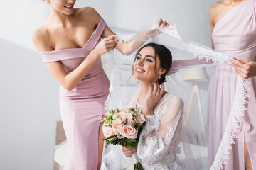 bridesmaids holding veil over pleased bride with wedding bouquet in bedroom.