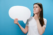 © Yura Yarema - Worried pensive confused young female holding empty speech bubble, spreading arms put hand prop up on chin, dressed in white t-shirt, isolated on blue studio background wall. Emotion people concept