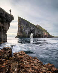 Person in front of Drangarnier rock formations on Vagar, water and seaweed in front, Faroe Islands.