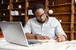 Focused African-American guy is using a laptop for watching webinars, taking notes, studying online. A male freelancer writes down startup ideas, sittin at the desk in office