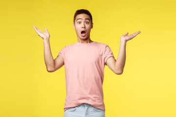 Vacation, summer and people concept. Portrait of surprised handsome, startled asian man gasping, raise hands sideways in amazement and awe, react to wonderful astonishing news, yellow background