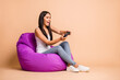 © deagreez - Full size profile photo of optimistic brunette girl sit pouf with playstation wear top jeans isolated on beige background