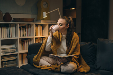 Wall Mural - young woman reading book while sitting on sofa at her home. she is covered with blanket and drinking coffee or tea