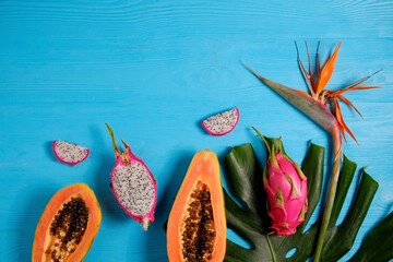 Wall Mural - Exotic fruit composition with monstera palm leaf. Papaya and pitaya, low-calorie nutritious fruits, whole and halved, visible pulp texture on blue wooden background. Close up, copy space, top view.