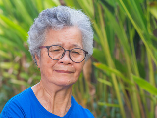 Portrait of a senior woman with short gray hair wearing glasses and looking down while standing in a garden. Space for text. Concept of aged people and healthcare