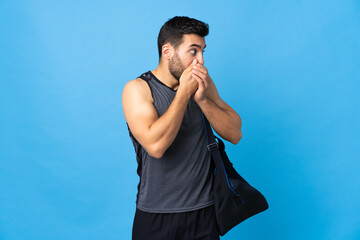 Wall Mural - Young sport man with sport bag isolated on blue background covering mouth and looking to the side