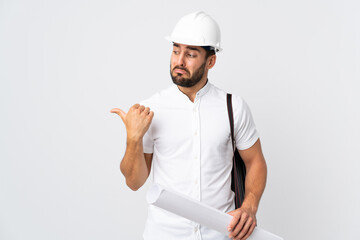 Canvas Print - Young architect man with helmet and holding blueprints isolated on white background unhappy and pointing to the side