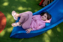 Cute Happy Beautiful Natural Teenage Girl In A Pink Jacket Is On A Playground With A Green Special Coating, Lies On A Children's Slide On A Sunny Day