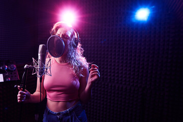 Wall Mural - Beautiful blonde girl emotionally singing song in recording studio with professional microphone and headphones, creates new track album, vocal artist in pink blue neon light