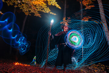 Sorcerer (magician) casting a spell while standing in the magical woods. Holding his magical staff. Magic illumination. Autumn holiday celebration. Mystery and nightmare concept.