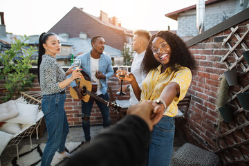 Wall Mural - Close up of male hand holding hand of smiling african woman with glass of champagne. Three happy friends singing and playing on guitar on background. Multiracial people on party.