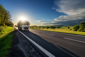 Wall Mural - white truck driving on the asphalt road in rural landscape in the rays of the sunset