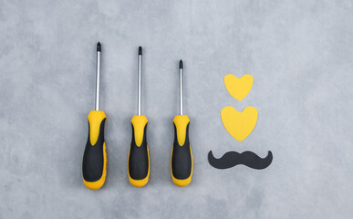 Wall Mural - A set of yellow screwdrivers with paper hearts and mustaches lie in the middle on a gray concrete background, close-up top view.