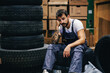 © dusanpetkovic1 - Young bored tattooed bearded worker in overalls sitting on tires in storage of import and export firm and waiting orders from his boss.