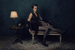 © ysbrandcosijn - Seductive young woman in retro boudoir attire with necklaces on an antique sofa next to a coffee table with a table lamp and a telephone.