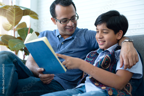 Indian father and son reading book at home together, son and father sit on couch at home laugh at funny joke