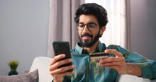 Close Up Portrait Of Hindu Happy Joyful Young Man In Eyeglasses Sits On Couch In Living Room In Modern Apartment Browsing On Smartphone Paying With Credit Card Buying Stuff Online, Shopping Concept