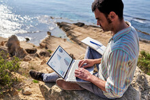 A young man works remotely with the sea in the background