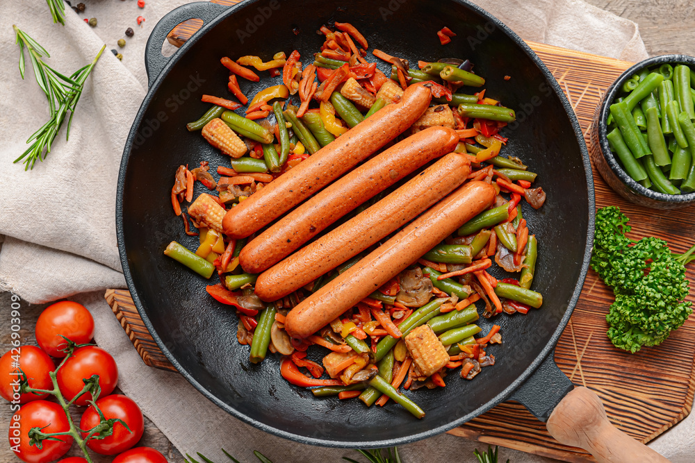 Frying pan with tasty sausages and vegetables on wooden background