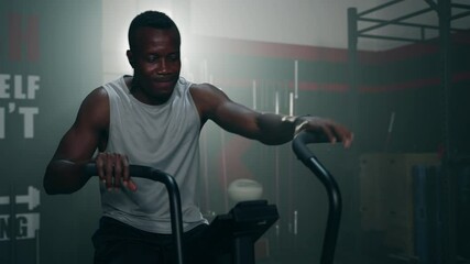 Wall Mural - African ethnicity man listening music while using spinning bike on cardio machine in fitness gym
