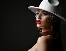 Portrait Of Rich Gorgeous Woman In Wide-brimmed White Hat With Veiling And Massive Gold Collar And Earrings. Stands With Bared Shoulders Over Black Background. Stylish Look And Sexual Games Concept