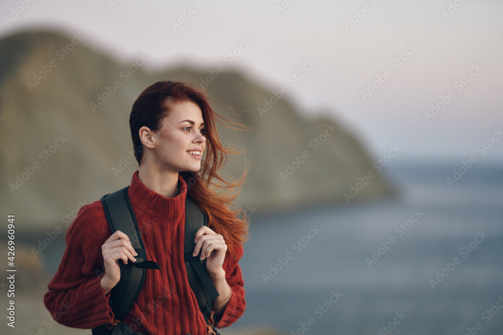 woman hiker holds a backpack on her shoulders and look at the sea from the side of the mountains in the background - obrazy, fototapety, plakaty 