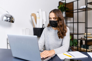 Wall Mural - Female employee stays in safety against viral diseases working in the office, woman wearing medical mask using the laptop indoor, protecting herself during pandemic