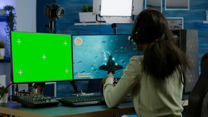 Wall Mural - African gamer playing virtual videogame on powerful computer, chatting on green screen mock up, chroma key display. Cyber player using pc with isolated desktop streaming shooter games wearing headset