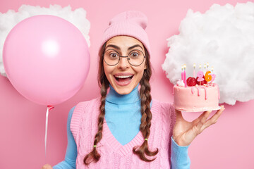 Happy beautiful woman celebrates birthday glad friends came to congratulate her poses with tasty cake and inflated balloon wears casual clothes poses against pink background. Festive event concept