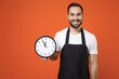 Young fun man 20s barista bartender barman employee in black apron white t-shirt work in coffee shop hold in hands clock isolated on orange background. Small business startup time management concept