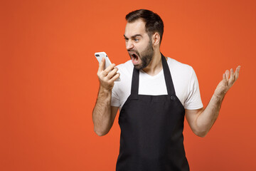 Wall Mural - Young angry man 20s barista bartender barman employee wear black apron white t-shirt work in coffee shop talk by mobile phone spread hands scream isolated on orange background. Small business startup.