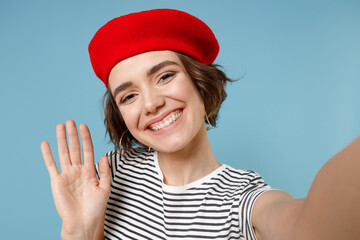 Wall Mural - Close up friendly young fun woman 20s with short hairdo in french beret red hat striped t-shirt do selfie shot on mobile phone waving hand greeting isolated on pastel blue background studio portrait.