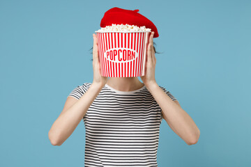 Wall Mural - Young scared woman 20s wearing french beret red hat striped t-shirt hiding covering face with popcorn takeaway bucket watching horror thriller isolated on pastel blue color background studio portrait.